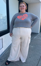 Load image into Gallery viewer, Full-body front view of a size 26 Eloquii off-white cargo-style wide leg trouser styled with a gray sweater and black slides on a size 22/4 model. The photo is taken outside in natural lighting.

