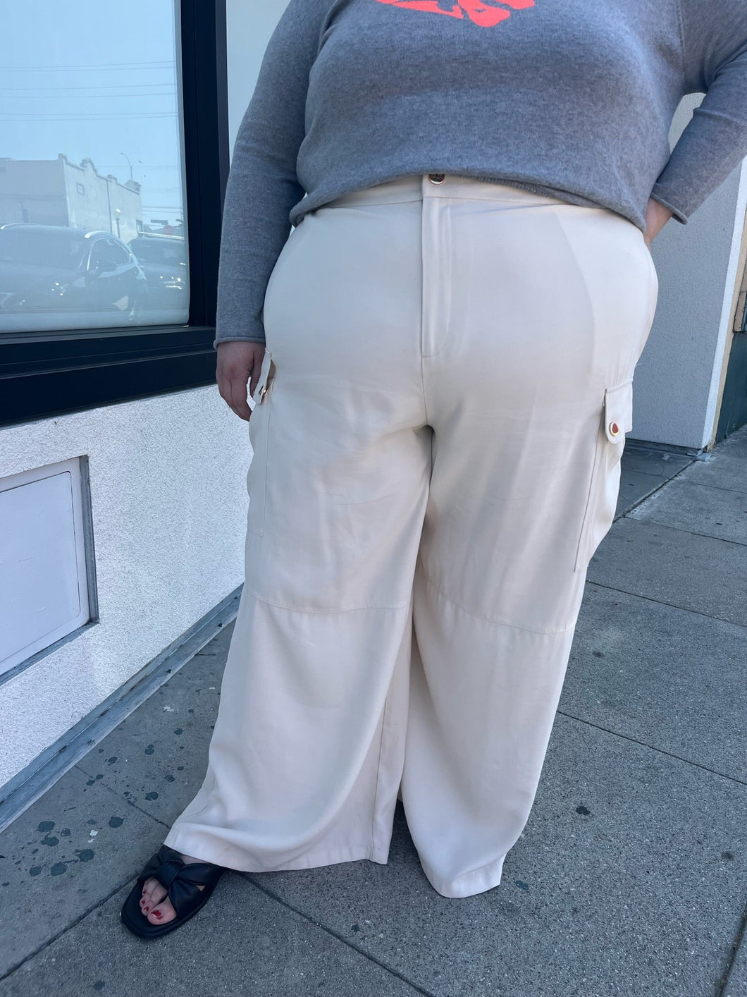 Front view of a size 26 Eloquii off-white cargo-style wide leg trouser styled with a gray sweater and black slides on a size 22/4 model. The photo is taken outside in natural lighting.