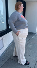 Load image into Gallery viewer, Full-body side view of a size 26 Eloquii off-white cargo-style wide leg trouser styled with a gray sweater and black slides on a size 22/4 model. The photo is taken outside in natural lighting.
