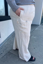 Load image into Gallery viewer, Side view of a size 26 Eloquii off-white cargo-style wide leg trouser styled with a gray sweater and black slides on a size 22/4 model. The photo is taken outside in natural lighting.
