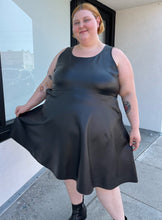 Load image into Gallery viewer, Final full-body front view of a size 4 Torrid black pleather tank-style a-line midi dress with pockets styled with black boots on a size 22/24 model. The photo is taken outside in natural lighting.
