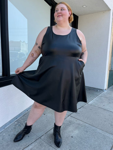 Full-body front view of a size 4 Torrid black pleather tank-style a-line midi dress with pockets styled with black boots on a size 22/24 model. The photo is taken outside in natural lighting.