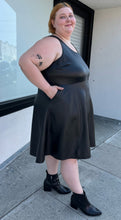 Load image into Gallery viewer, Full-body side view of a size 4 Torrid black pleather tank-style a-line midi dress with pockets styled with black boots on a size 22/24 model. The photo is taken outside in natural lighting.
