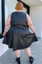 Load image into Gallery viewer, Full-body back view of a size 4 Torrid black pleather tank-style a-line midi dress with pockets styled with black boots on a size 22/24 model. The photo is taken outside in natural lighting.
