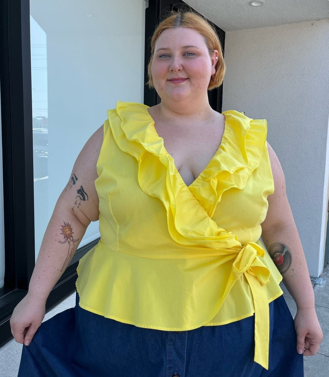 Front view of a size 4 Fashion to Figure x Gabrielle Union collab bright yellow ruffled wrap blouse styled with a denim skirt on a size 22/24 model. The photo is taken outside in natural lighting.