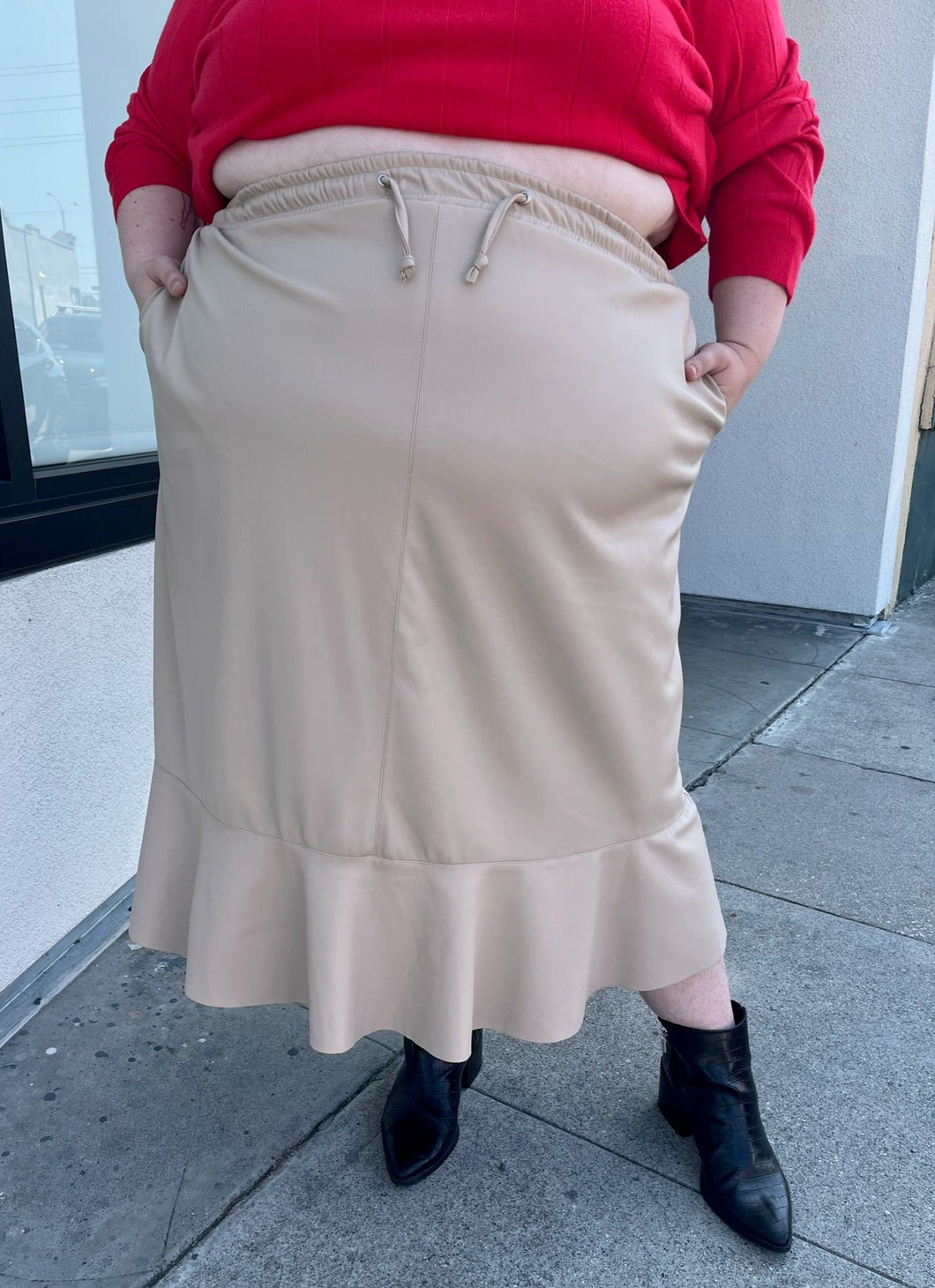 Front view of a size 24/26 Ulla Popken khaki maxi skirt with drawstring waist and ruffle hem styled with a red sweater and black boots on a size 22/24 model. The photo is taken outside in natural lighting.