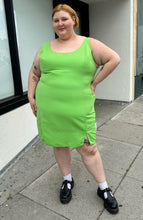 Load image into Gallery viewer, Full-body front view of a size 24 ASOS lime green mini sheath dress with a small slit at the hem styled with black mary janes on a size 22/24 model. The photo is taken outside in natural lighting.
