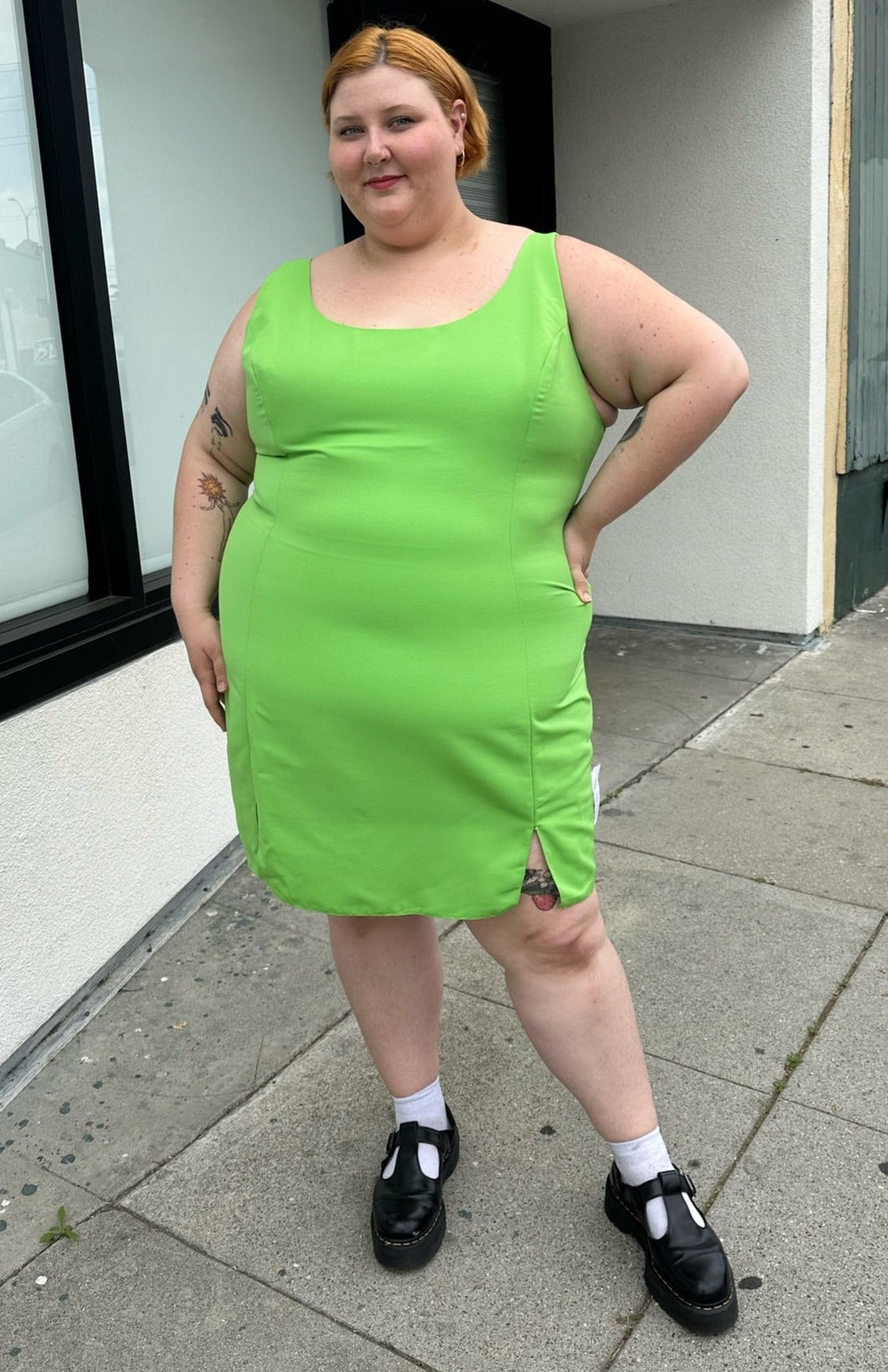 Full-body front view of a size 24 ASOS lime green mini sheath dress with a small slit at the hem styled with black mary janes on a size 22/24 model. The photo is taken outside in natural lighting.