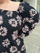 Load image into Gallery viewer, Closer view of the off-shoulder details of a size 24 Collusion black off-shoulder tent dress with sketched pink daisies, a ruffle hem, and ruffle cuffs on a size 22/24 model. The photo is taken outside in natural lighting.
