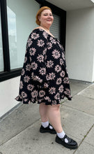 Load image into Gallery viewer, Full-body side view of a size 24 Collusion black off-shoulder tent dress with sketched pink daisies, a ruffle hem, and ruffle cuffs styled with black mary janes on a size 22/24 model. The photo is taken outside in natural lighting.
