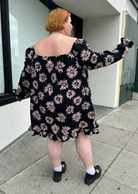 Load image into Gallery viewer, Full-body back view of a size 24 Collusion black off-shoulder tent dress with sketched pink daisies, a ruffle hem, and ruffle cuffs styled with black mary janes on a size 22/24 model. The photo is taken outside in natural lighting.
