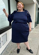 Load image into Gallery viewer, Additional full-body front view of a size 22 Eloquii navy blue midi dress with ruffle details and tie-detail cold shoulders styled with black slides on a size 22/24 model. The photo is taken outside in natural lighting.
