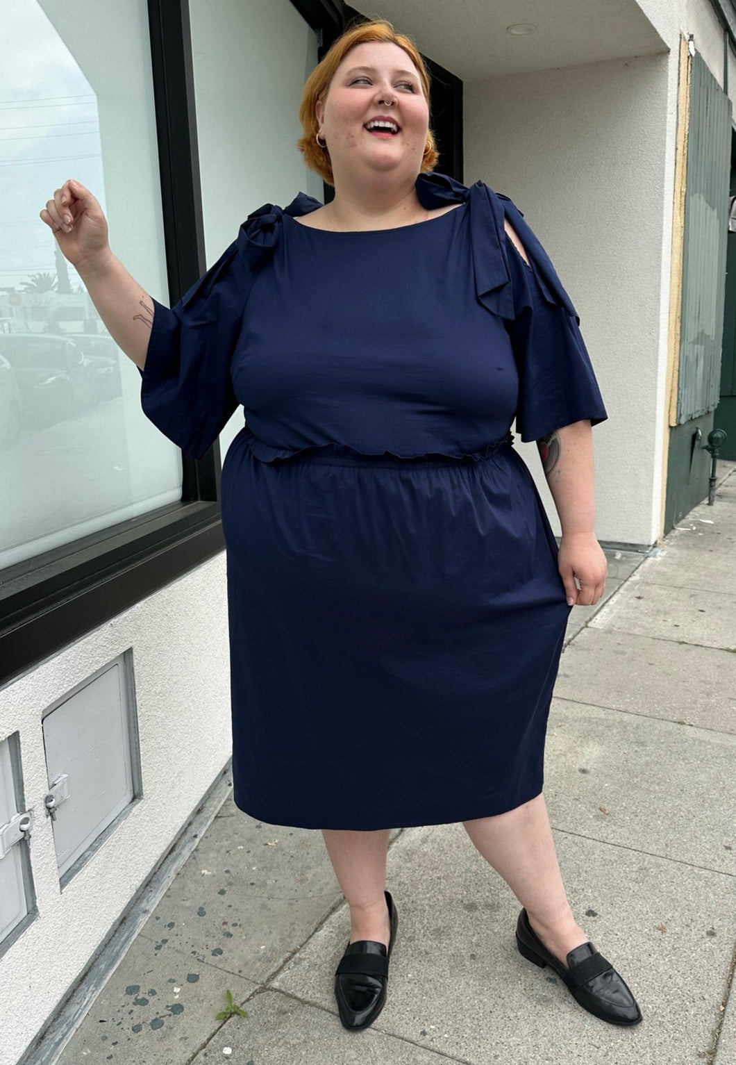 Full-body front view of a size 22 Eloquii navy blue midi dress with ruffle details and tie-detail cold shoulders styled with black slides on a size 22/24 model. The photo is taken outside in natural lighting.