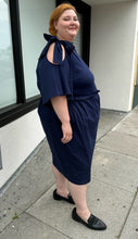 Load image into Gallery viewer, Full-body side view of a size 22 Eloquii navy blue midi dress with ruffle details and tie-detail cold shoulders styled with black slides on a size 22/24 model. The photo is taken outside in natural lighting.
