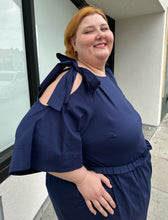Load image into Gallery viewer, Closer side view of the tie-detail cold shoulder of a size 22 Eloquii navy blue midi dress with ruffle details and tie-detail cold shoulders on a size 22/24 model. The photo is taken outside in natural lighting.
