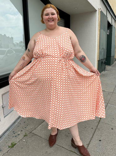 Full-body front view of a size 5X WRAY NYC orange and white checkered windowpane tank maxi dress with tie belt styled with brown mules on a size 22/24 model. The photo is taken outside in natural lighting.