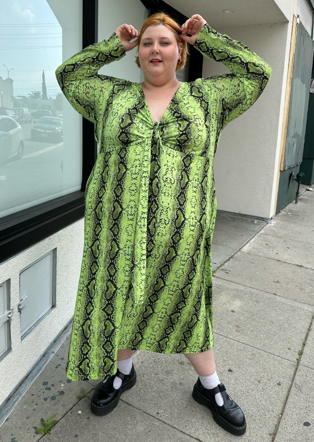 Full-body front view of a size 26 ASOS lime green and black snake patterned maxi with twist-front tie detail and long sleeves styled with black mary janes on a size 22/24 model.. The photo is taken outside in natural lighting.