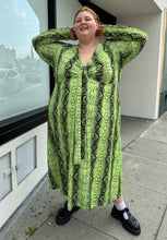 Load image into Gallery viewer, Final full-body front view of a size 26 ASOS lime green and black snake patterned maxi with twist-front tie detail and long sleeves styled with black mary janes on a size 22/24 model.. The photo is taken outside in natural lighting.
