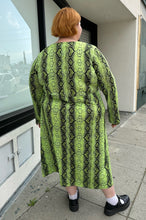 Load image into Gallery viewer, Full-body back view of a size 26 ASOS lime green and black snake patterned maxi with twist-front tie detail and long sleeves styled with black mary janes on a size 22/24 model.. The photo is taken outside in natural lighting.
