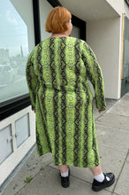 Load image into Gallery viewer, Full-body back view of a size 26 ASOS lime green and black snake patterned maxi with twist-front tie detail and long sleeves styled with black mary janes on a size 22/24 model.. The photo is taken outside in natural lighting.
