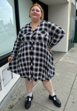 Load image into Gallery viewer, Additional full-body front view of a size 3 Torrid black and white plaid collarless a-line shirt dress with breast pockets styled with black mary janes on a size 22/24 model. The photo is taken outside in natural lighting.
