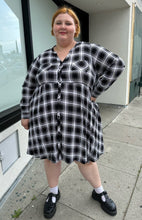 Load image into Gallery viewer, Full-body front view of a size 3 Torrid black and white plaid collarless a-line shirt dress with breast pockets styled with black mary janes on a size 22/24 model. The photo is taken outside in natural lighting.
