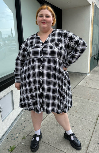 Full-body front view of a size 3 Torrid black and white plaid collarless a-line shirt dress with breast pockets styled with black mary janes on a size 22/24 model. The photo is taken outside in natural lighting.