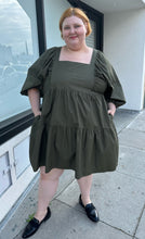 Load image into Gallery viewer, Full-body front view showing off the pockets of a size 3X A New Day olive green square neck puff sleeve tiered dress styled with black slides on a size 22/24 model. The photo is taken outside in natural lighting.
