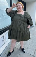 Load image into Gallery viewer, Full-body front view of a size 3X A New Day olive green square neck puff sleeve tiered dress styled with black slides on a size 22/24 model. The photo is taken outside in natural lighting.
