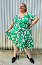 Load image into Gallery viewer, Additional full-body front view of a size 22 Maggie London green and white abstract pattern wrap dress with asymmetrical hemline styled with black slip on loafers on a size 22/24 model. The photo is taken outside in natural lighting.

