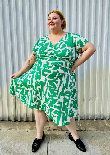 Full-body front view of a size 22 Maggie London green and white abstract pattern wrap dress with asymmetrical hemline styled with black slip on loafers on a size 22/24 model. The photo is taken outside in natural lighting.
