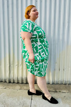 Load image into Gallery viewer, Full-body side view of a size 22 Maggie London green and white abstract pattern wrap dress with asymmetrical hemline styled with black slip on loafers on a size 22/24 model. The photo is taken outside in natural lighting.
