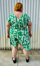 Load image into Gallery viewer, Full-body back view of a size 22 Maggie London green and white abstract pattern wrap dress with asymmetrical hemline styled with black slip on loafers on a size 22/24 model. The photo is taken outside in natural lighting.
