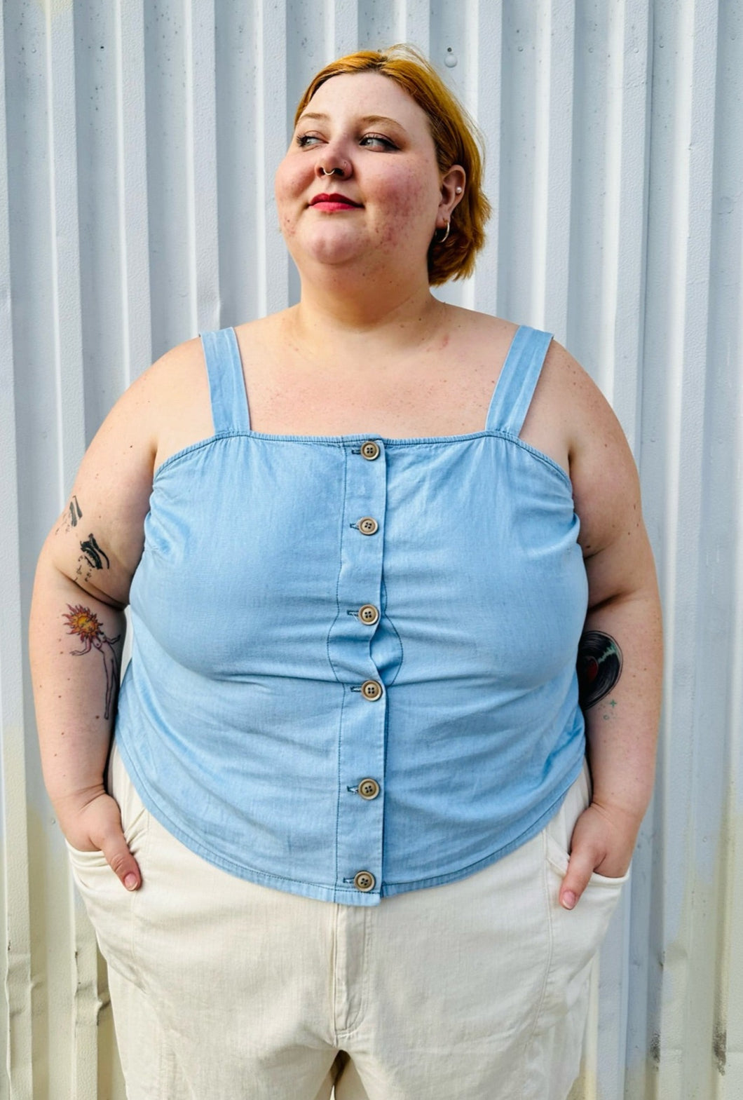 Front view of a size 24 Lane Bryant light blue button-up tank with brown buttons, thick straps, and a smocked back styled with cream shorts on a size 22/24 model. The photo is taken outside in natural lighting.
