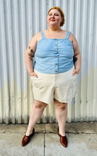 Load image into Gallery viewer, Full-body front view of a size 24 Lane Bryant light blue button-up tank with brown buttons, thick straps, and a smocked back styled with cream shorts and brown mules on a size 22/24 model. The photo is taken outside in natural lighting.
