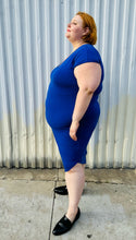 Load image into Gallery viewer, Full-body side view of a size M (Universal Standard size 18/20) royal blue v-neck t-shirt dress styled with black loafer slides on a size 22/24 model. The photo is taken outside in natural lighting.
