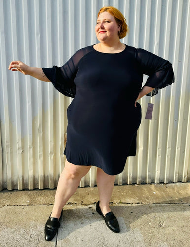 Full-body front view of a size 2X Nina Leonard navy blue mini dress with sheer mesh tiered sleeves styled with black slide loafers. The photo is taken outside in natural lighting.