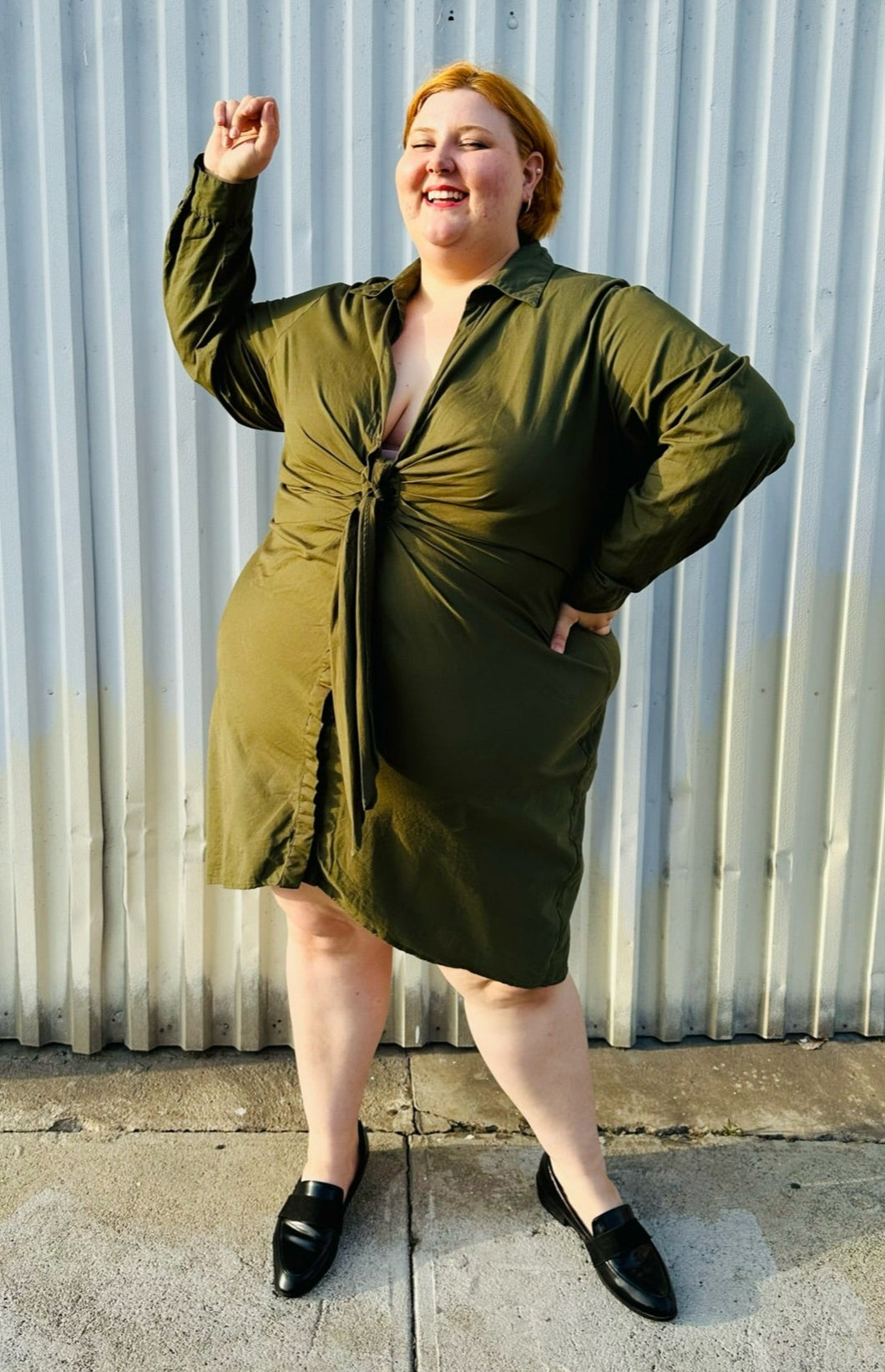 Full-body front view of a size 4 Fashion to Figure army green twist-front collared shirt dress styled with black slide loafers on a size 22/24 model. The photo is taken outside in natural lighting.