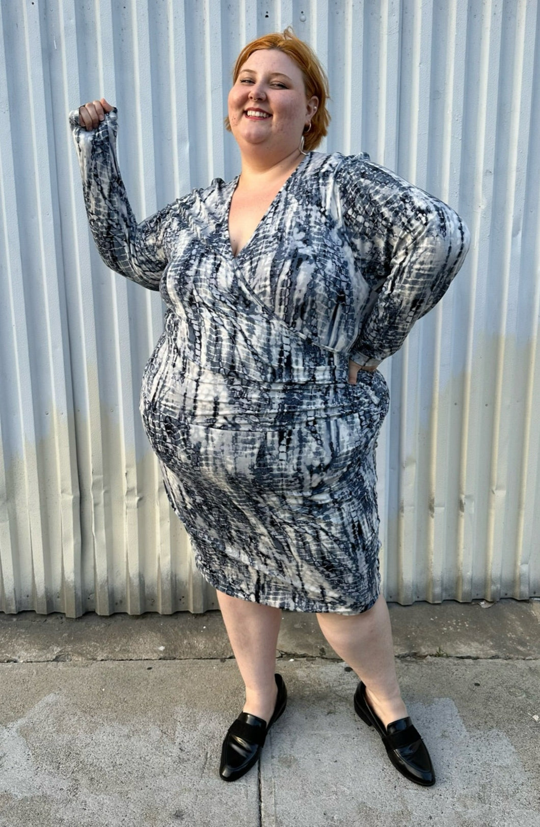 Full-body front view showing off the long sleeves of a size 4X Tart black, blue, and white abstract tie dye long sleeve faux wrap dress styled with black loafer slides on a size 22/24 model. The photo is taken outside in natural lighting.