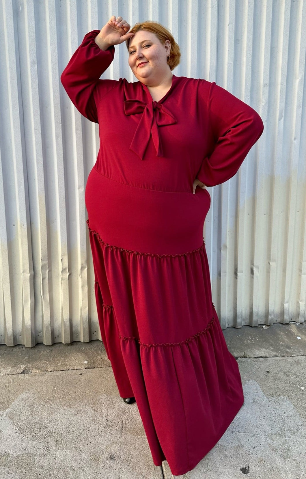 Full-body front view of a size 3X Melissa Masse Made to Measure deep red tiered maxi dress with subtle long puff sleeves, pussy bow neckline, and long tiers on a size 22/24 model. The photo is taken outside in natural lighting.