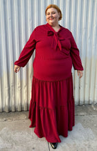 Load image into Gallery viewer, Additional full-body front view of a size 3X Melissa Masse Made to Measure deep red tiered maxi dress with subtle long puff sleeves, pussy bow neckline, and long tiers on a size 22/24 model. The photo is taken outside in natural lighting.
