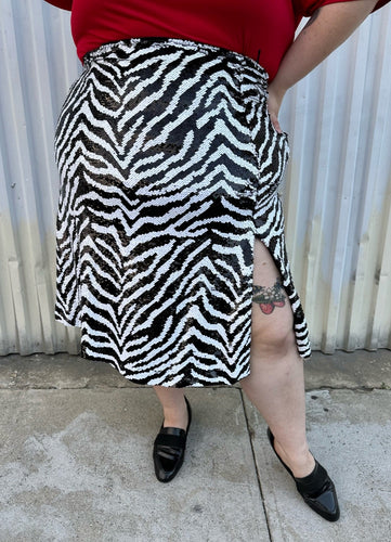 Front view of a size 22 Novella black and white sequin zebra pattern maxi skirt with side slit styled with a red top and black loafer slides on a size 22/24 model. The photo is taken outside in natural lighting.