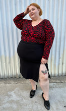 Load image into Gallery viewer, Full-body front view of a size 22/24 Eloquii black and red leopard pattern long sleeve bodysuit styled tucked into a black high-slit skirt and black loafer slides on a size 22/24 model. The photo is taken outside in natural lighting.
