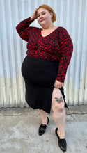 Load image into Gallery viewer, Full-body front view of a size 26 Lane Bryant black stretchy midi skirt with convertible high side slit styled with a red &amp; black leopard bodysuit and black loafers on a size 22/24 model. The photo is taken outside in natural lighting.
