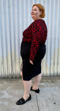 Load image into Gallery viewer, Full-body side view of a size 26 Lane Bryant black stretchy midi skirt with convertible high side slit styled with a red &amp; black leopard bodysuit and black loafers on a size 22/24 model. The photo is taken outside in natural lighting.
