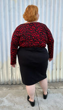 Load image into Gallery viewer, Full-body back view of a size 26 Lane Bryant black stretchy midi skirt with convertible high side slit styled with a red &amp; black leopard bodysuit and black loafers on a size 22/24 model. The photo is taken outside in natural lighting.
