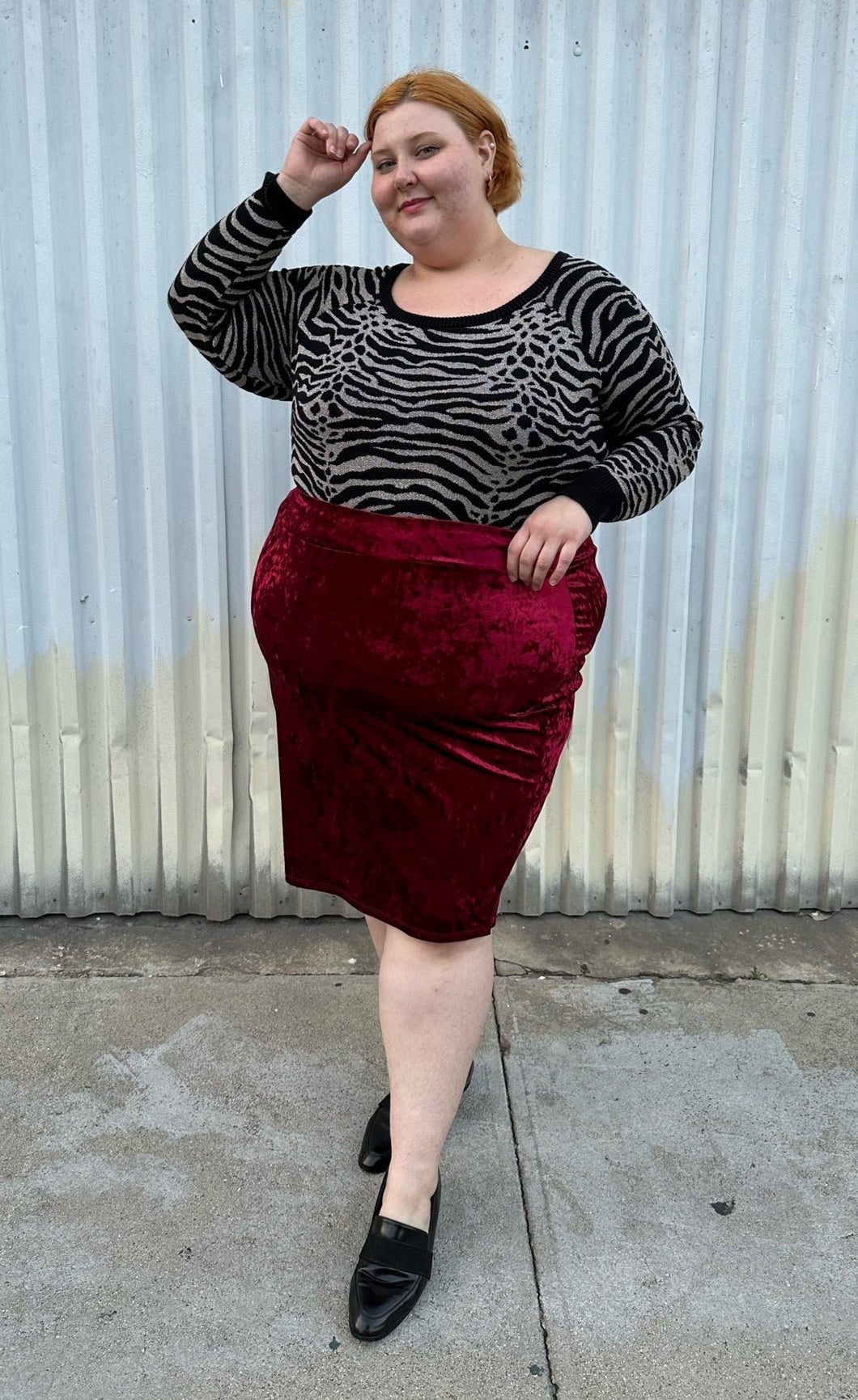 Full-body front view of a size 5X SWAK deep red crushed velvet bodycon midi pencil skirt styled with an animal print sweater tucked in and loafers on a size 22/24 model. The photo is taken outside in natural lighting.