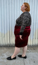 Load image into Gallery viewer, Full-body side view of a size 5X SWAK deep red crushed velvet bodycon midi pencil skirt styled with an animal print sweater tucked in and loafers on a size 22/24 model. The photo is taken outside in natural lighting.
