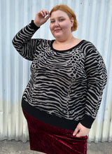 Load image into Gallery viewer, Front view of a size 3 Torrid black and silver metallic thread animal pattern sweater with black piping and cuffs styled with a red crushed velvet bodycon skirt on a size 22/24 model. The photo is taken outside in natural lighting.
