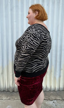 Load image into Gallery viewer, Side view of a size 3 Torrid black and silver metallic thread animal pattern sweater with black piping and cuffs styled with a red crushed velvet bodycon skirt on a size 22/24 model. The photo is taken outside in natural lighting.
